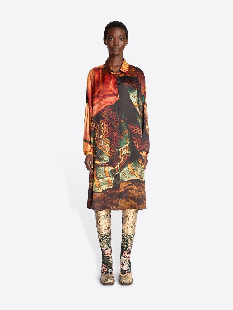 A model stands against a light gray background wearing a long-sleeve collared shirt dress with an intricate multicolor abstract print , paired with patterned tights and snakeskin heels.