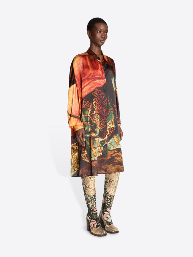 A model stands against a light gray background wearing a long-sleeve collared shirt dress with an intricate multicolor abstract print , paired with patterned tights and snakeskin heels.