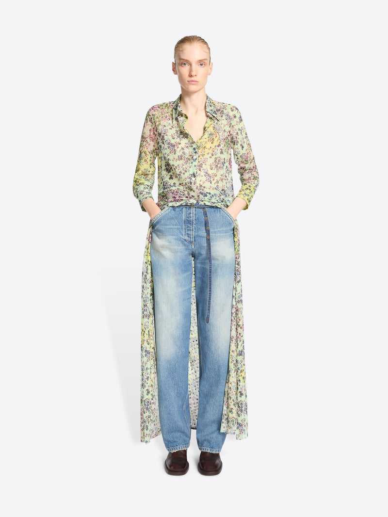 A model stands against a light gray background wearing a long-sleeve button-front shirt with a dense multicolor floral print, paired with light-wash blue jeans and dark brown shoes.