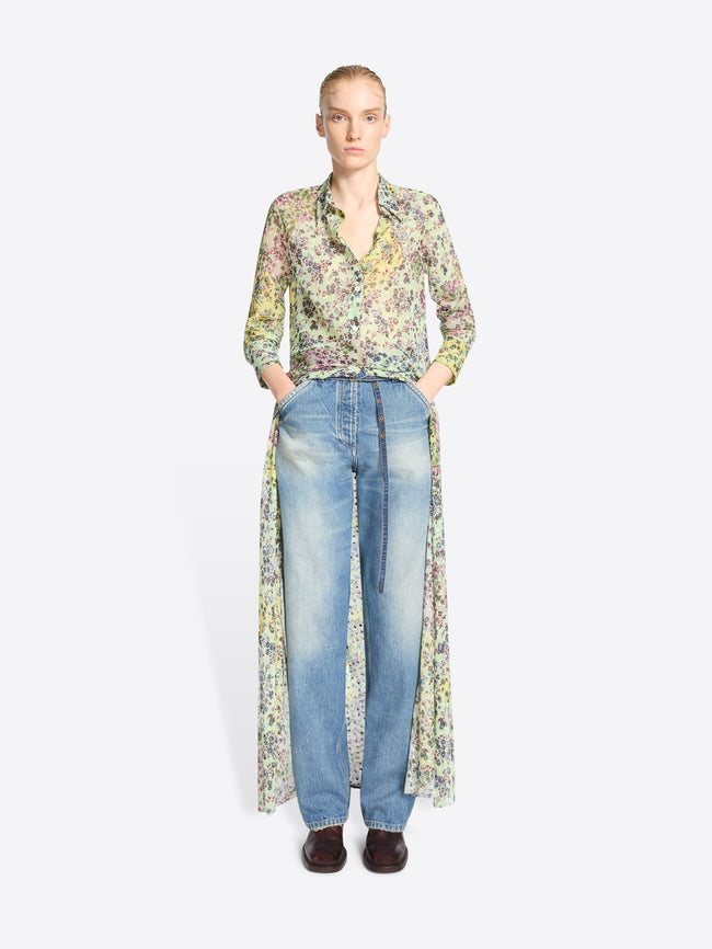 A model stands against a light gray background wearing a long-sleeve button-front shirt with a dense multicolor floral print, paired with light-wash blue jeans and dark brown shoes.