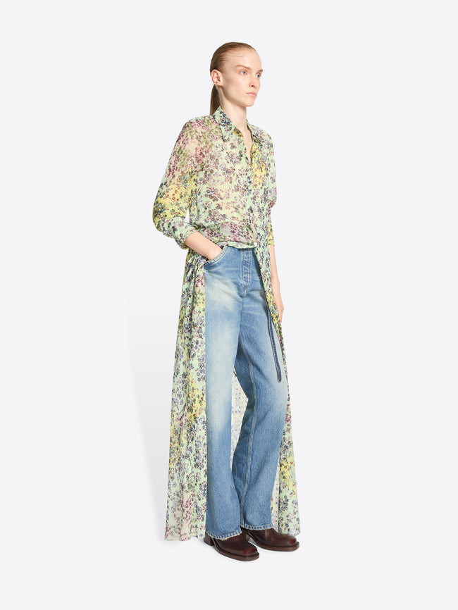 A model stands against a light gray background wearing a long-sleeve button-front shirt with a dense multicolor floral print, paired with light-wash blue jeans and dark brown shoes.