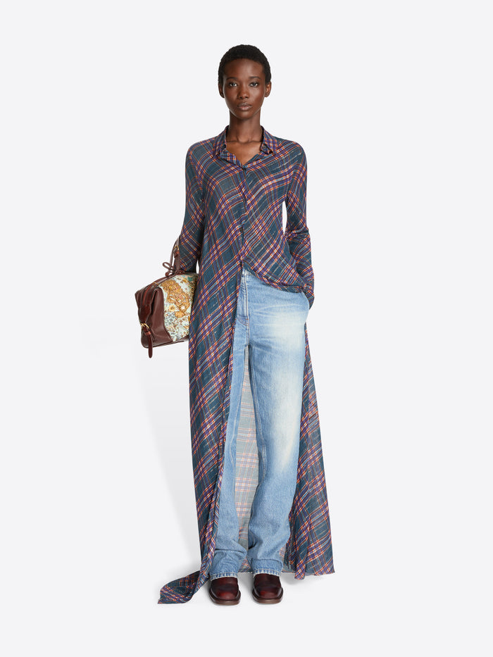A model stands against a light gray background wearing a long-sleeve button-front shirt dress with a pink and blue plaid pattern over light blue denim jeans, holding a small brown handbag.