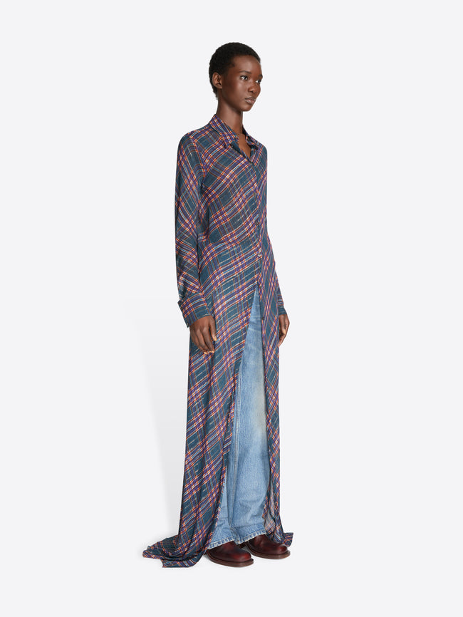 A model stands against a light gray background wearing a long-sleeve button-front shirt dress with a pink and blue plaid pattern over light blue denim jeans.
