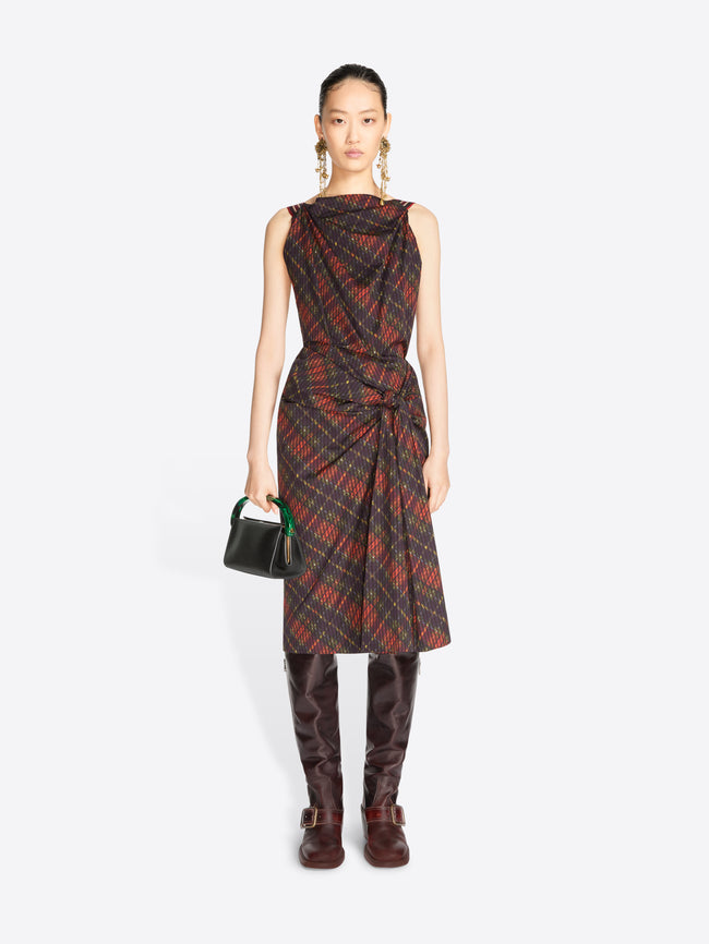 A model stands against a light gray background wearing a sleeveless draped midi dress with a red and black plaid pattern, paired with dark brown knee-high boots and holding a small black handbag.