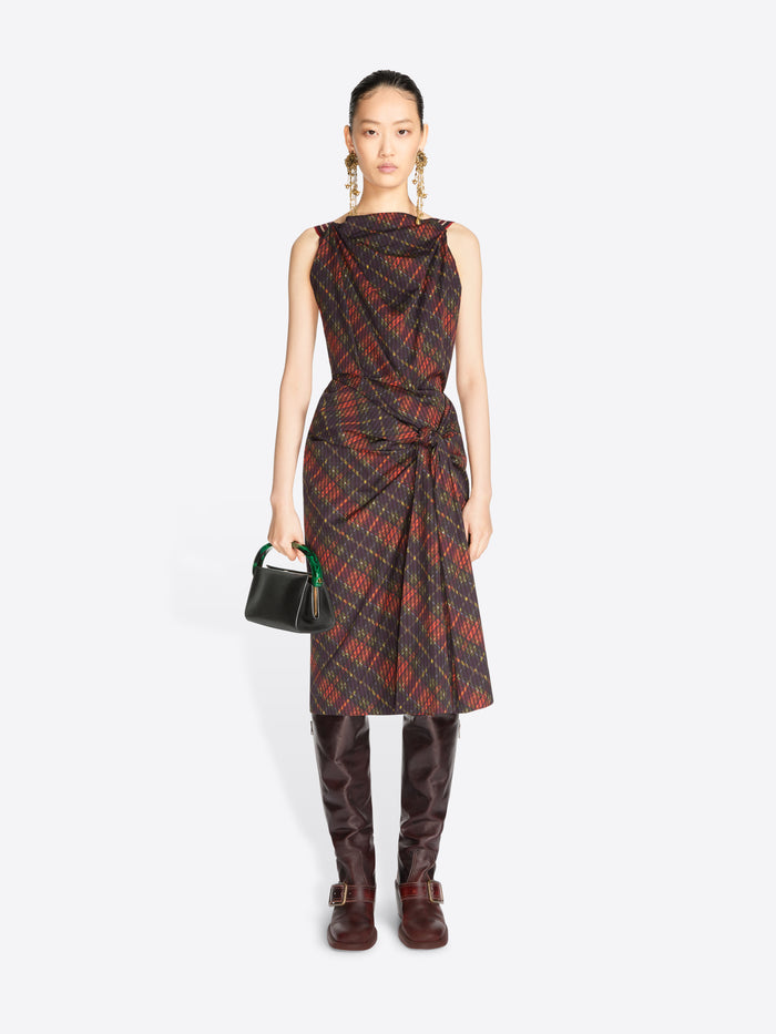 A model stands against a light gray background wearing a sleeveless draped midi dress with a red and black plaid pattern, paired with dark brown knee-high boots and holding a small black handbag.