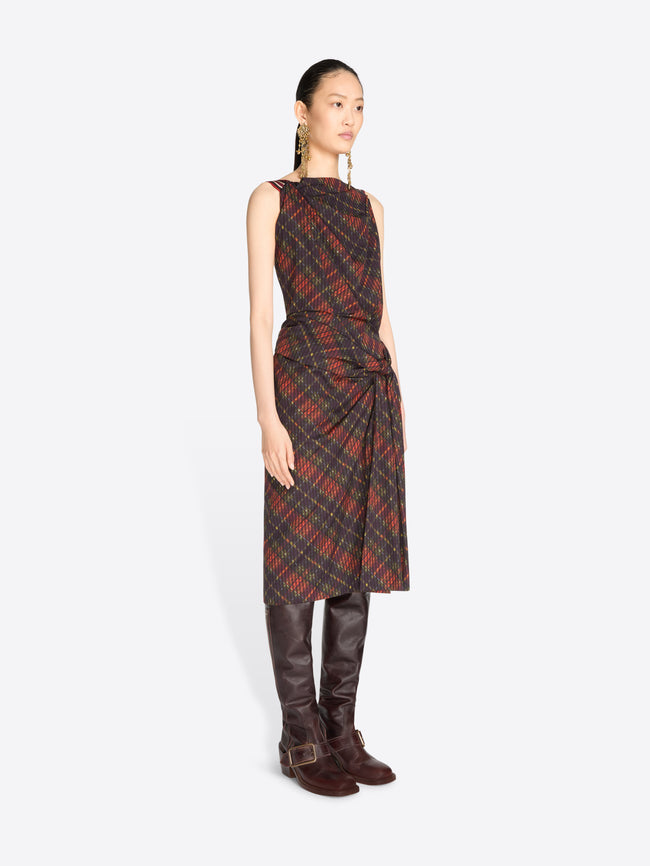 A model stands against a light gray background wearing a sleeveless draped midi dress with a red and black plaid pattern, paired with dark brown knee-high boots.