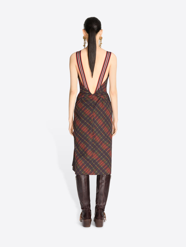 Back view of a model stands against a light gray background wearing a sleeveless draped midi dress with a red and black plaid pattern, paired with dark brown knee-high boots.