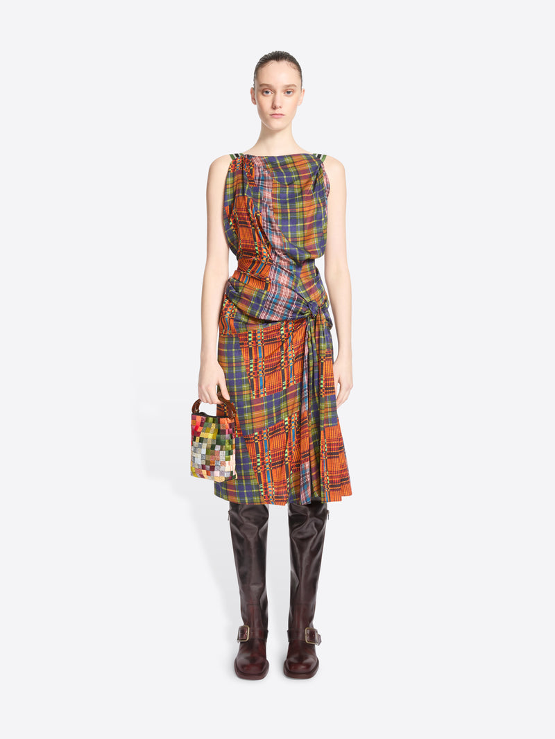 A model stands against a light gray background wearing a sleeveless wrap-style dress with a multicolor plaid pattern paired with dark brown knee-high boots and holding a small multicolor handbag.
