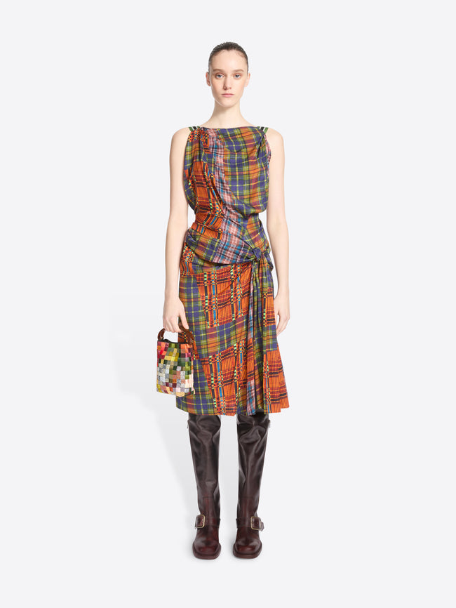 A model stands against a light gray background wearing a sleeveless wrap-style dress with a multicolor plaid pattern paired with dark brown knee-high boots and holding a small multicolor handbag.