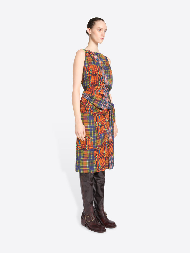 A model stands against a light gray background wearing a sleeveless wrap-style dress with a multicolor plaid pattern paired with dark brown knee-high boots.