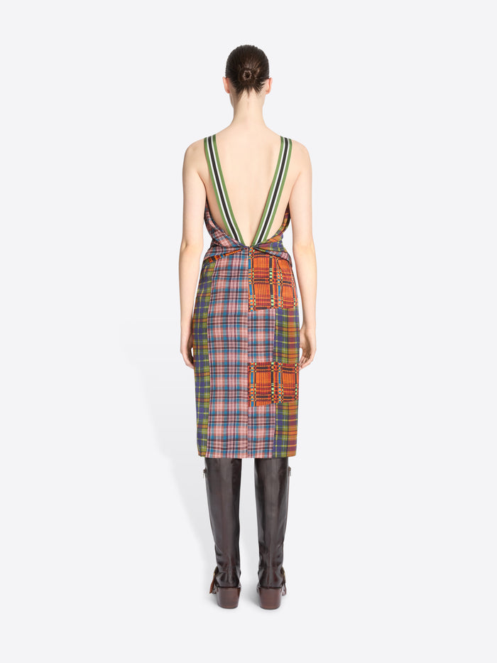 Back view of a model wearing a sleeveless multicolor plaid with patchwork details dress with a deep V-back, striped shoulder straps, paired with brown knee-high boots against a light gray background.