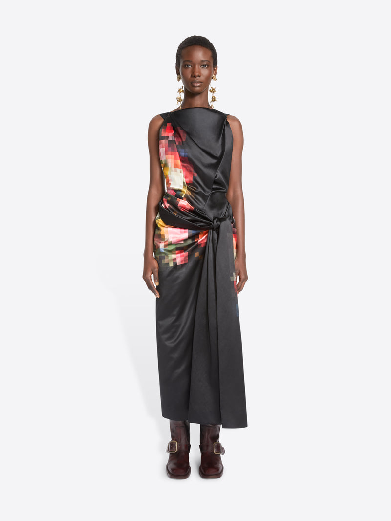 A model stands against a light gray background wearing a sleeveless black wrap-style dress with a colorful abstract print on the upper back and side, paired with dark brown ankle boots.