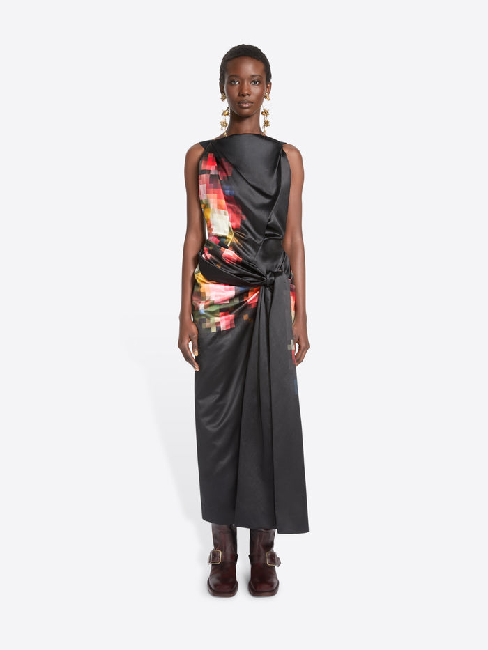 A model stands against a light gray background wearing a sleeveless black wrap-style dress with a colorful abstract print on the upper back and side, paired with dark brown ankle boots.