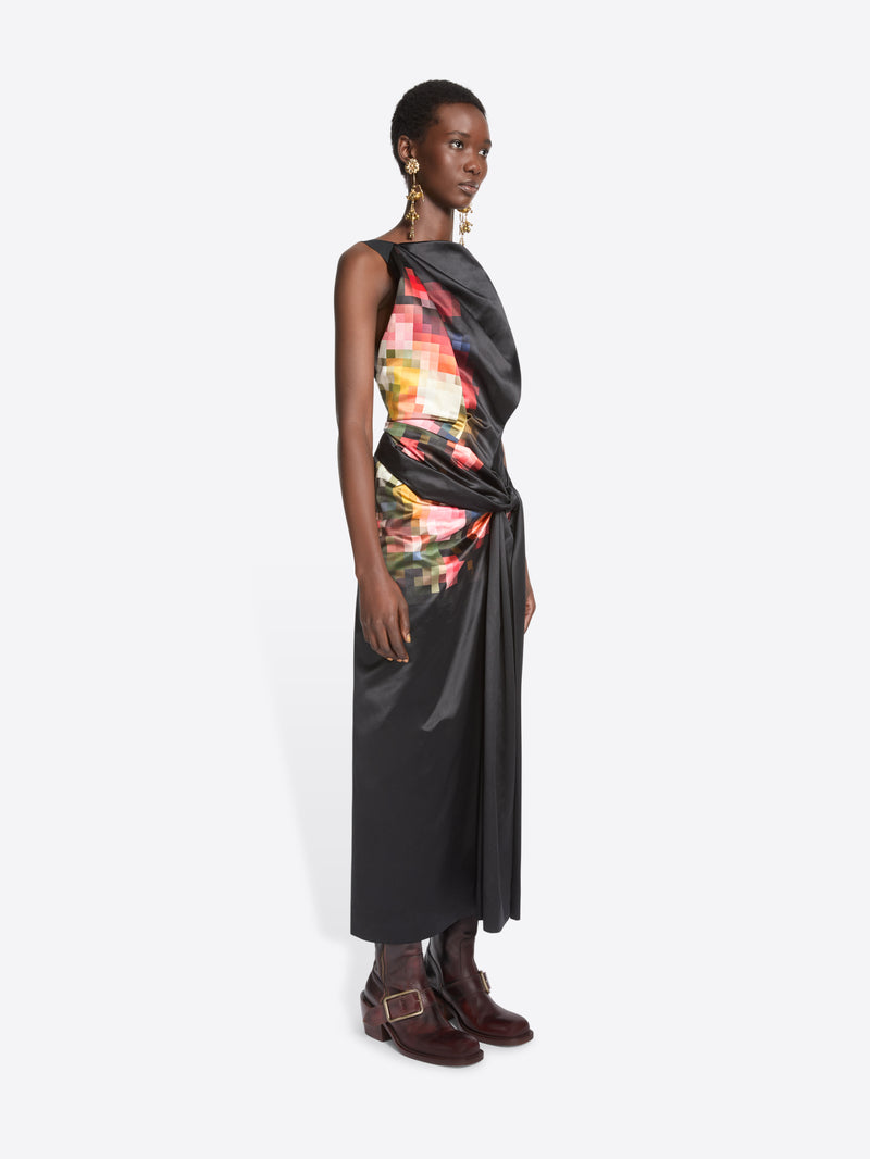 A model stands against a light gray background wearing a sleeveless black wrap-style dress with a colorful abstract print on the upper back and side, paired with dark brown ankle boots.