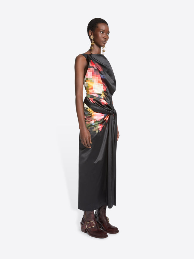 A model stands against a light gray background wearing a sleeveless black wrap-style dress with a colorful abstract print on the upper back and side, paired with dark brown ankle boots.