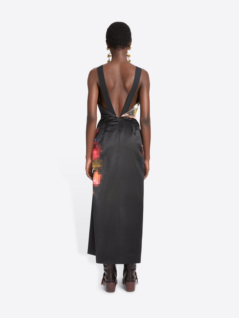 Back view of a model standing against a light gray background wearing a sleeveless black wrap-style dress with a colorful abstract print on the upper back and side, paired with dark brown ankle boots.