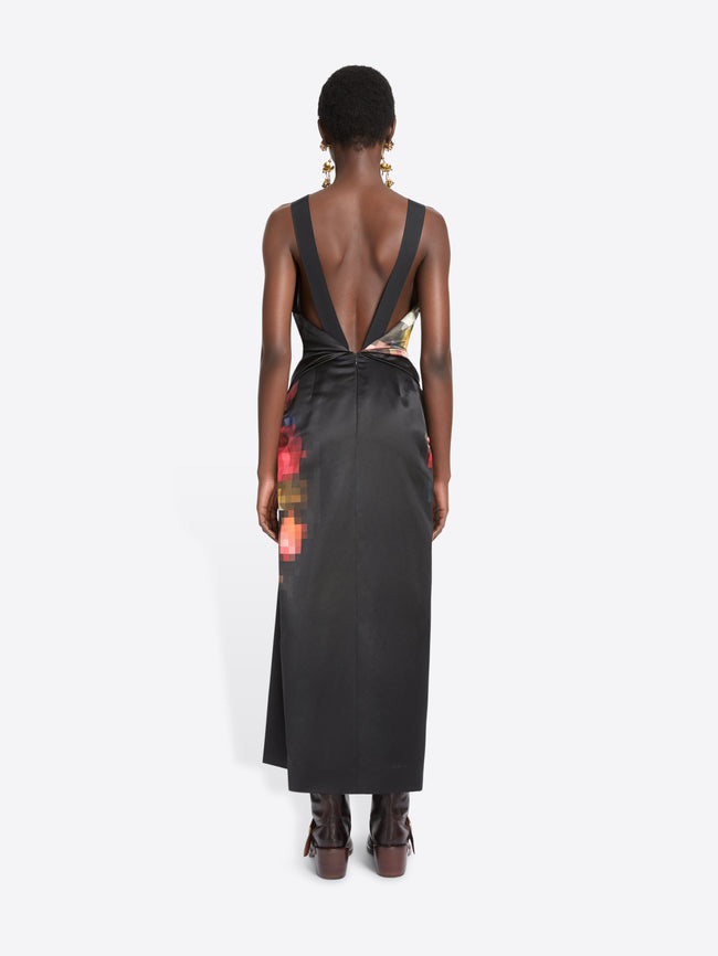 Back view of a model standing against a light gray background wearing a sleeveless black wrap-style dress with a colorful abstract print on the upper back and side, paired with dark brown ankle boots.