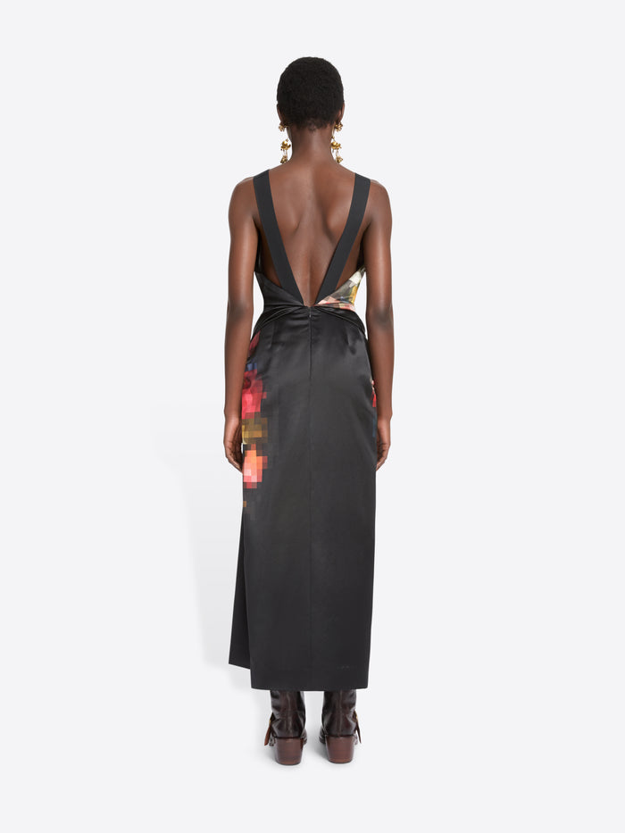 Back view of a model standing against a light gray background wearing a sleeveless black wrap-style dress with a colorful abstract print on the upper back and side, paired with dark brown ankle boots.