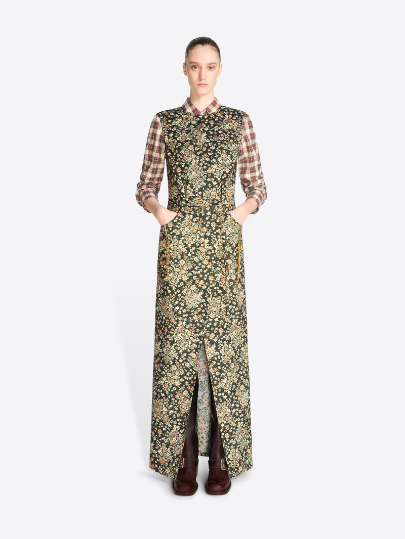 A model stands against a light gray background wearing a long-sleeved maxi dress with a dense floral print in olive green and beige, layered over a brown and beige plaid shirt, paired with dark brown boots.