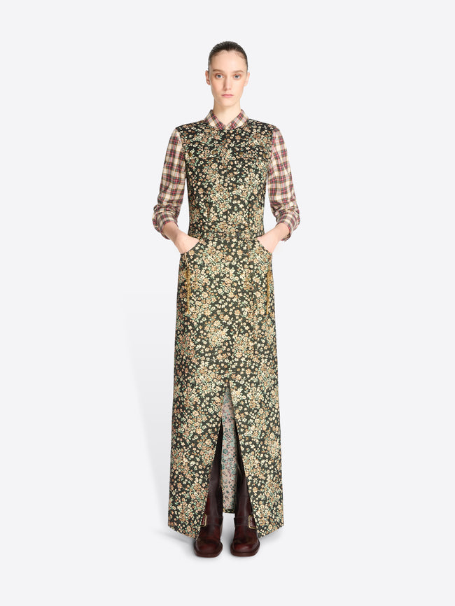 A model stands against a light gray background wearing a long-sleeved maxi dress with a dense floral print in olive green and beige, layered over a brown and beige plaid shirt, paired with dark brown boots.