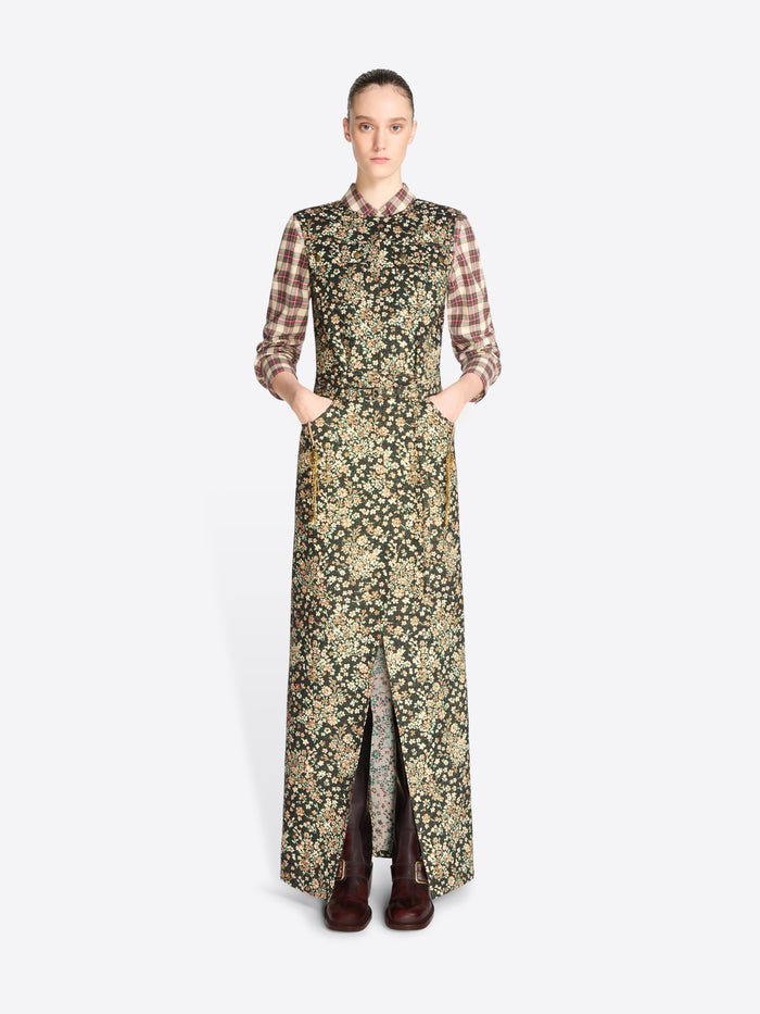 A model stands against a light gray background wearing a long-sleeved maxi dress with a dense floral print in olive green and beige, layered over a brown and beige plaid shirt, paired with dark brown boots.