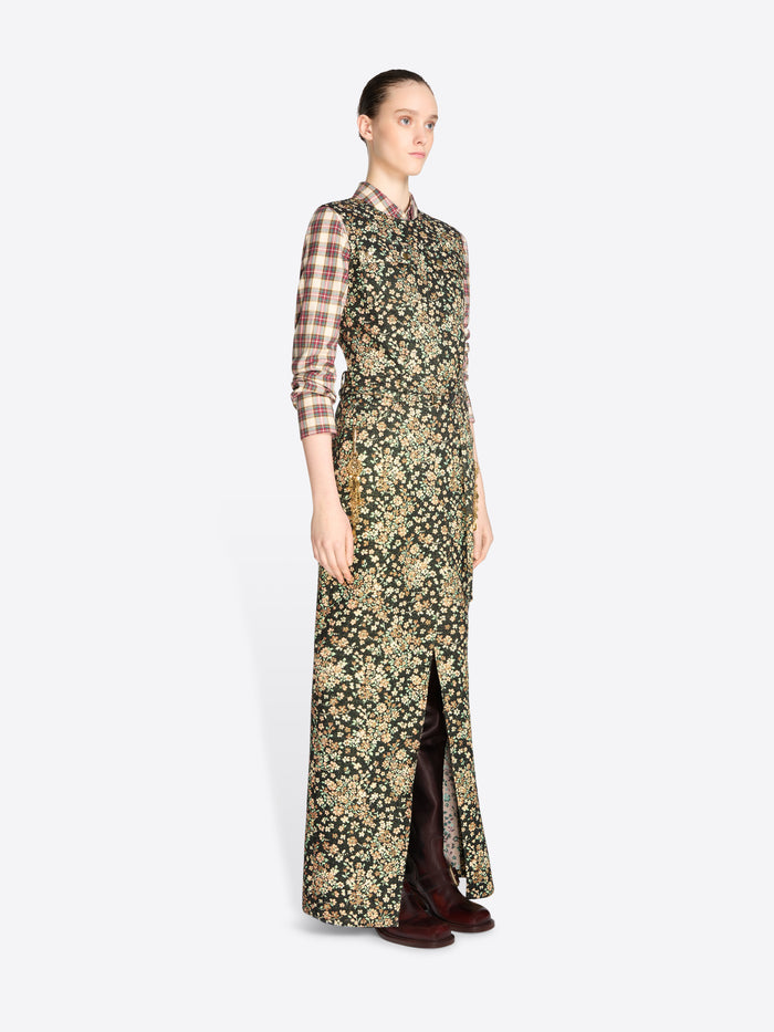 A model stands against a light gray background wearing a long-sleeved maxi dress with a dense floral print in olive green and beige, layered over a brown and beige plaid shirt, paired with dark brown boots.