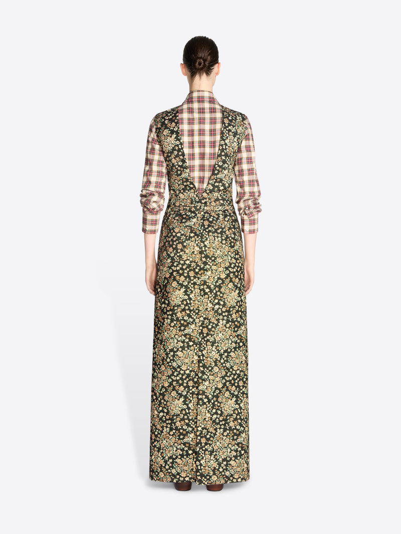 Back view of a model standing against a light gray background wearing a long-sleeved maxi dress with a dense floral print in olive green and beige, layered over a brown and beige plaid shirt, paired with dark brown boots.