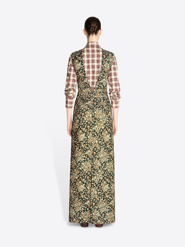 Back view of a model standing against a light gray background wearing a long-sleeved maxi dress with a dense floral print in olive green and beige, layered over a brown and beige plaid shirt, paired with dark brown boots.