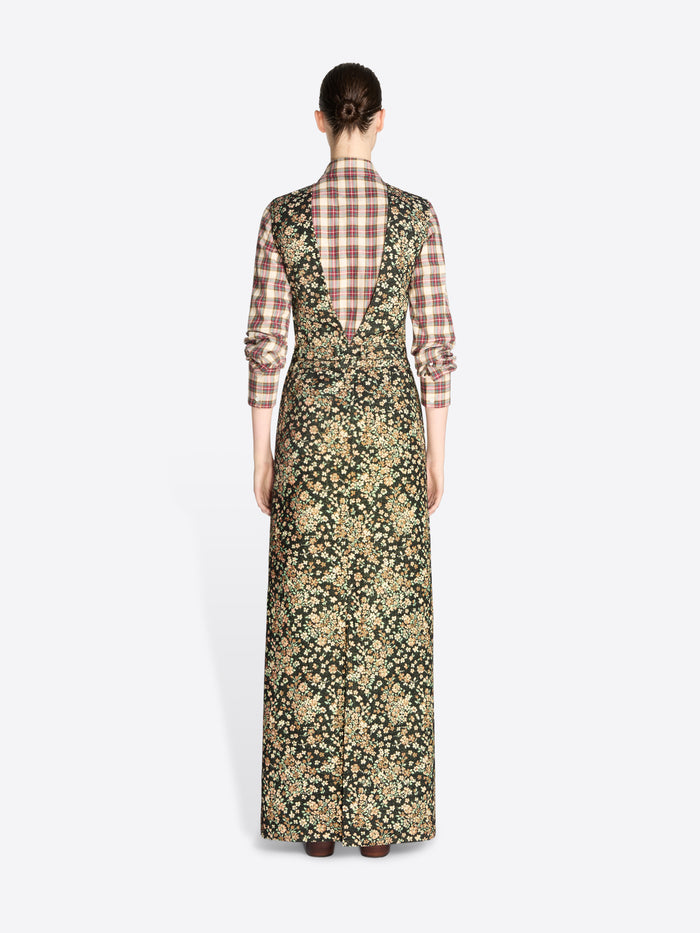 Back view of a model standing against a light gray background wearing a long-sleeved maxi dress with a dense floral print in olive green and beige, layered over a brown and beige plaid shirt, paired with dark brown boots.