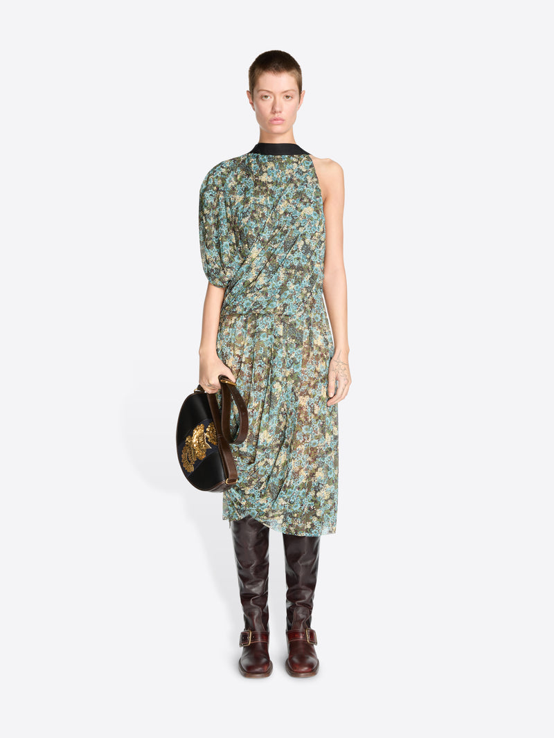 A model stands against a light gray background wearing a sleeveless, one-shoulder midi dress with a green and blue floral print, paired with dark brown knee-high boots and holding a black handbag.