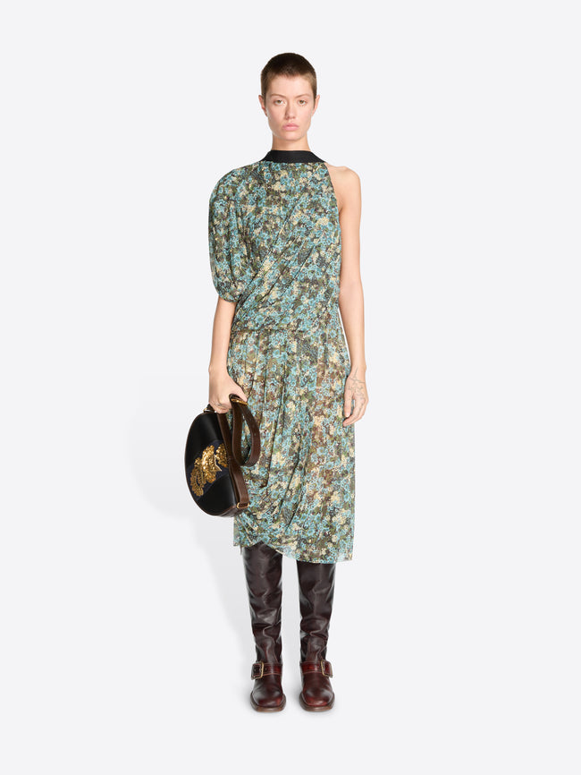 A model stands against a light gray background wearing a sleeveless, one-shoulder midi dress with a green and blue floral print, paired with dark brown knee-high boots and holding a black handbag.