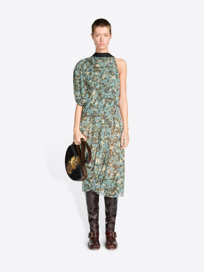 A model stands against a light gray background wearing a sleeveless, one-shoulder midi dress with a green and blue floral print, paired with dark brown knee-high boots and holding a black handbag.