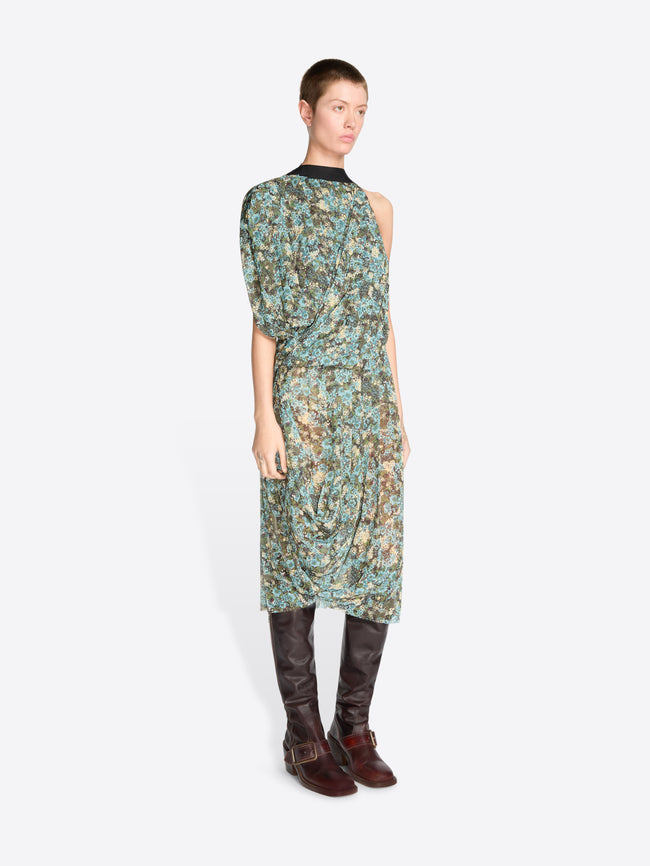 A model stands against a light gray background wearing a sleeveless, one-shoulder midi dress with a green and blue floral print, paired with dark brown knee-high boots.