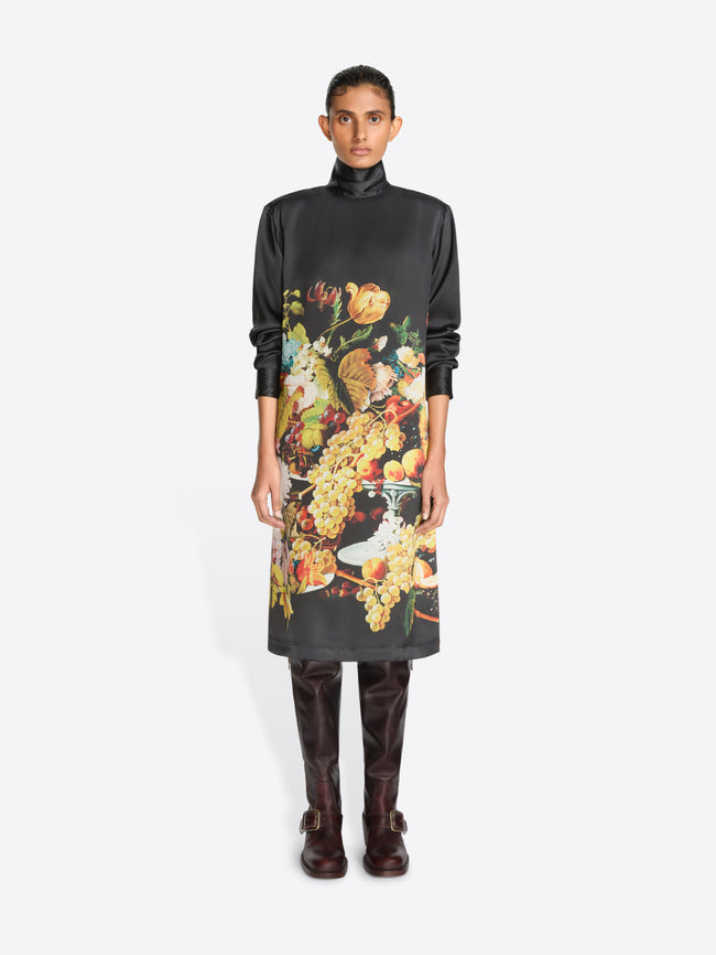 A model stands against a light gray background wearing a black turtleneck dress with a large, colorful floral and fruit print on the front, paired with dark brown knee-high boots.