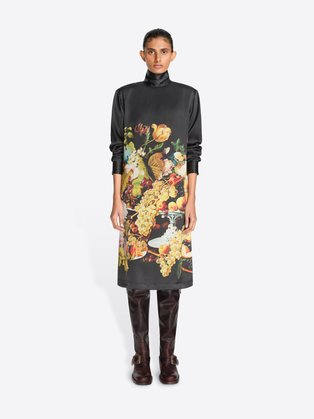A model stands against a light gray background wearing a black turtleneck dress with a large, colorful floral and fruit print on the front, paired with dark brown knee-high boots.