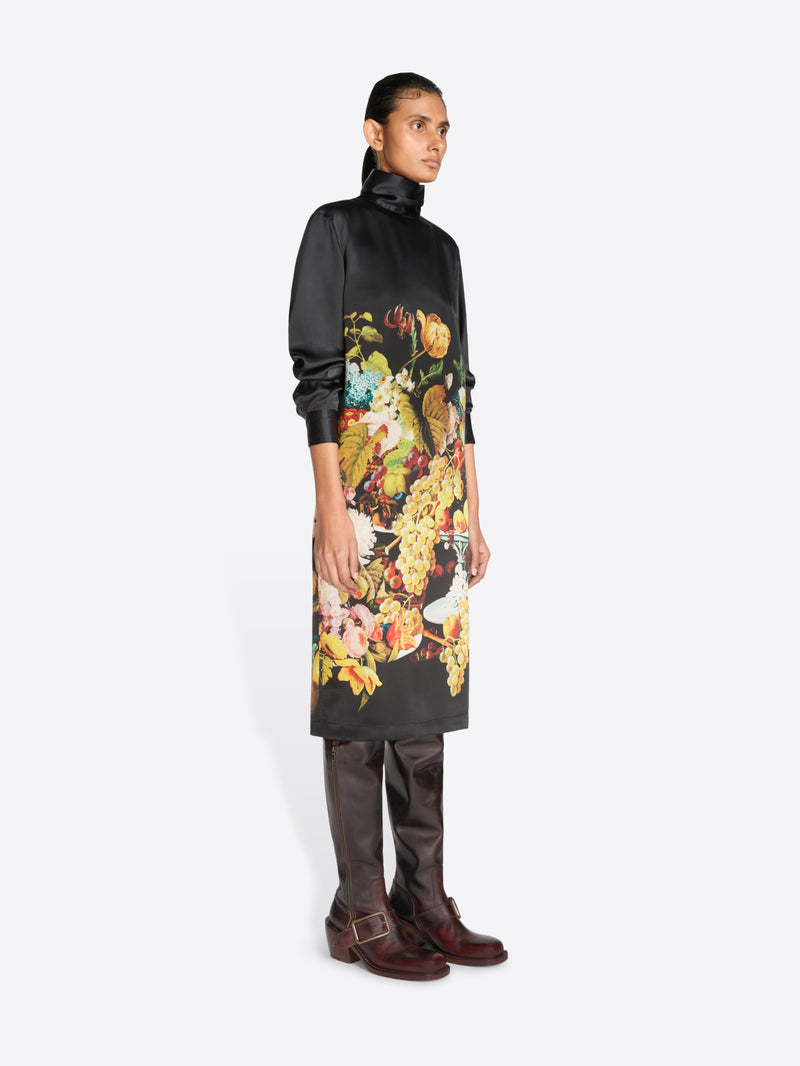 A model stands against a light gray background wearing a black turtleneck dress with a large, colorful floral and fruit print on the front, paired with dark brown knee-high boots.