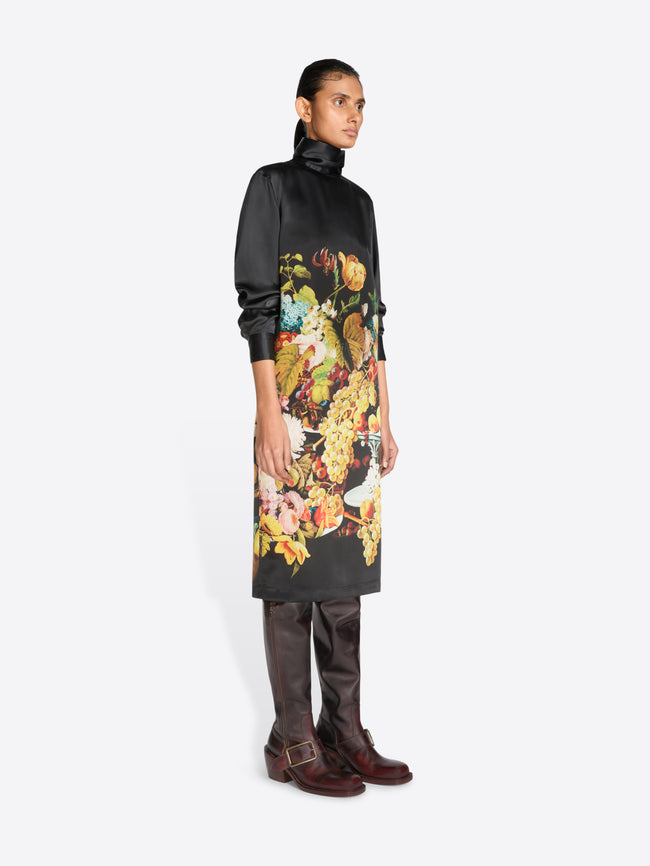 A model stands against a light gray background wearing a black turtleneck dress with a large, colorful floral and fruit print on the front, paired with dark brown knee-high boots.