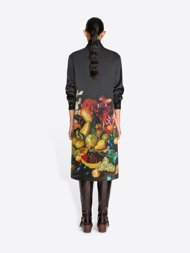 Back view of a model standing against a light gray background wearing a black turtleneck dress with a large, colorful floral and fruit print on the back, paired with dark brown knee-high boots.