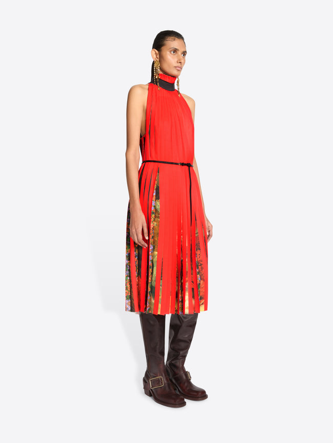 A model stands against a light gray background wearing a sleeveless high-neck red dress with a pleated skirt and a thin black belt, paired with dark brown knee-high boots.