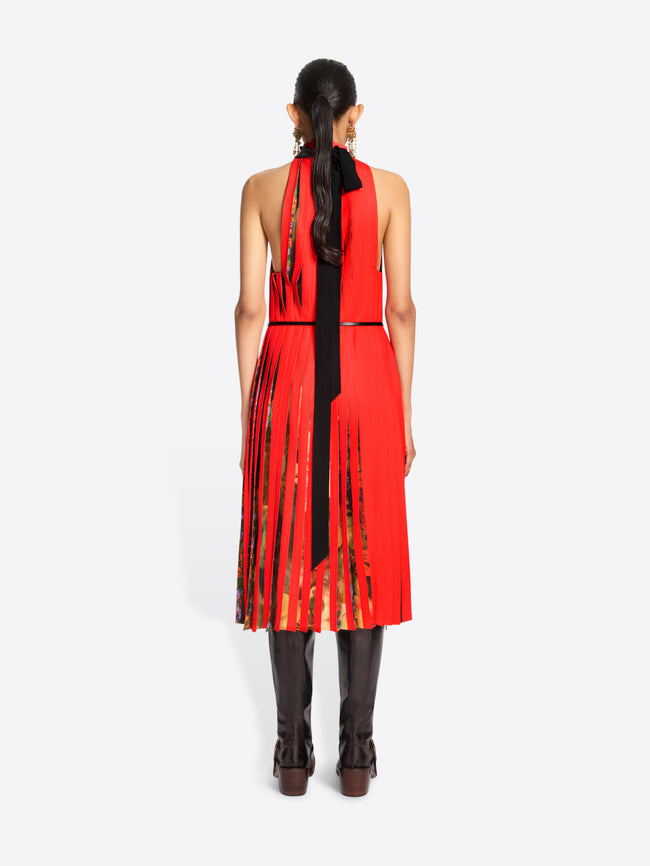 Back view of  a model standing against a light gray background wearing a sleeveless high-neck red dress with a pleated skirt and a thin black belt, paired with dark brown knee-high boots.