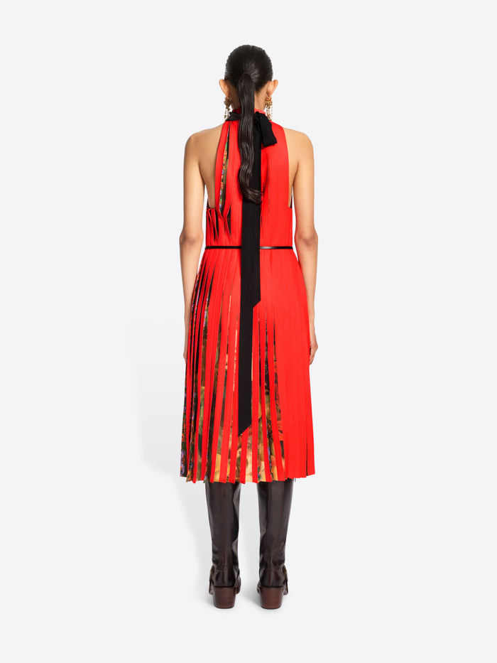 Back view of  a model standing against a light gray background wearing a sleeveless high-neck red dress with a pleated skirt and a thin black belt, paired with dark brown knee-high boots.