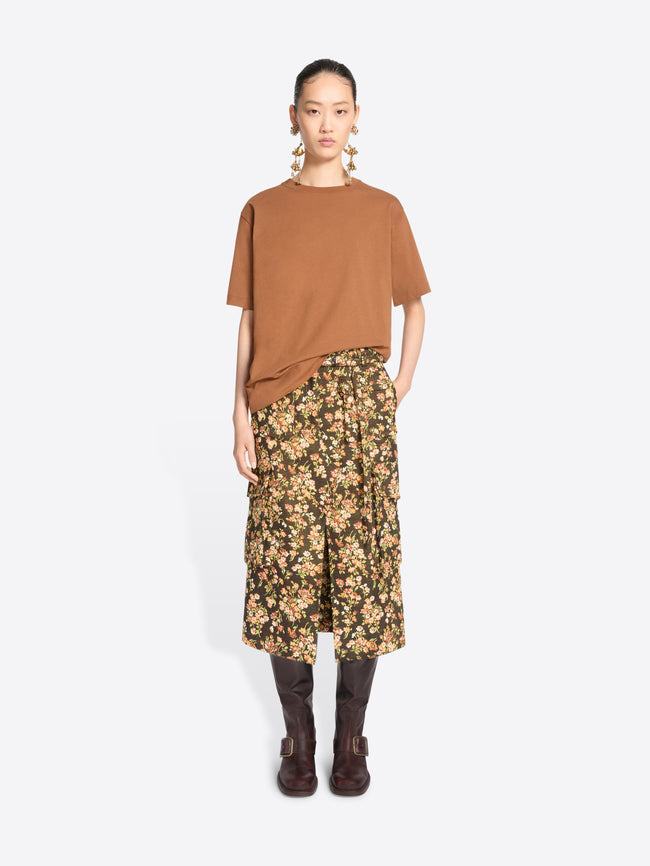 A model stands against a light gray background wearing a loose short-sleeve t-shirt, a dark olive floral midi skirt with yellow and beige flowers, and dark brown knee-high boots.