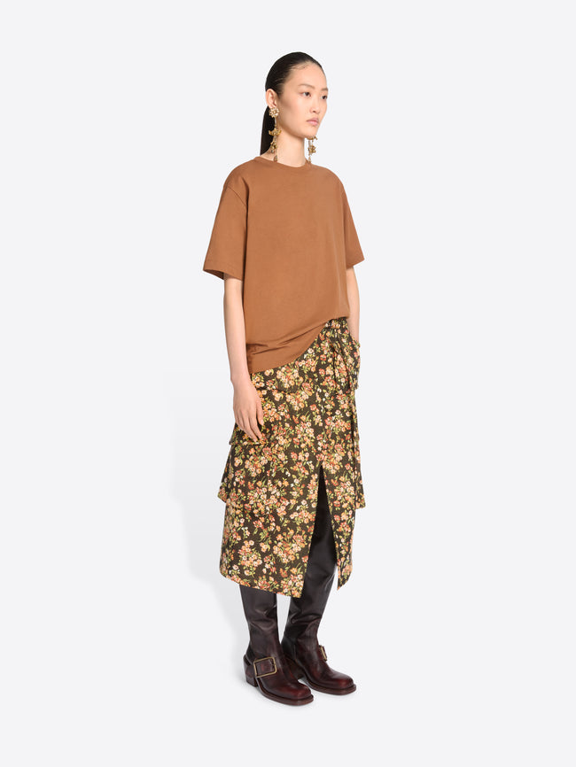 A model stands against a light gray background wearing a loose short-sleeve t-shirt, a dark olive floral midi skirt with yellow and beige flowers, and dark brown knee-high boots.