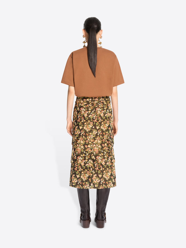 Back view of a model standing against a light gray background wearing a loose short-sleeve t-shirt, a dark olive floral midi skirt with yellow and beige flowers, and dark brown knee-high boots.