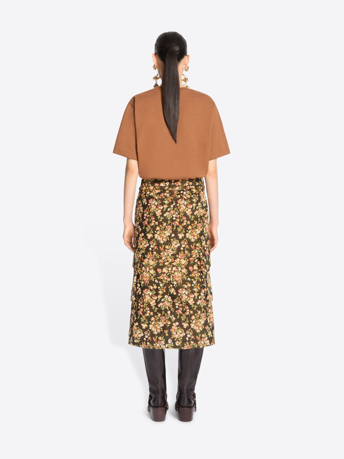 Back view of a model standing against a light gray background wearing a loose short-sleeve t-shirt, a dark olive floral midi skirt with yellow and beige flowers, and dark brown knee-high boots.