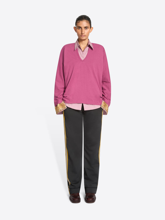 A model stands against a light gray background wearing a pink long-sleeve sweater over a light pink collared shirt, paired with dark gray track pants featuring yellow side stripes and dark brown shoes.