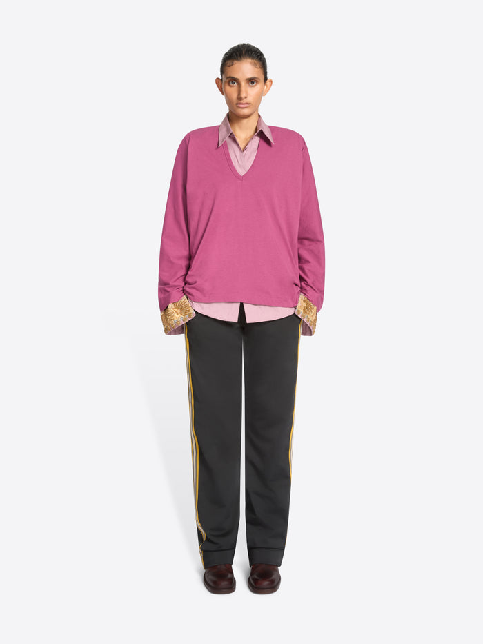 A model stands against a light gray background wearing a pink long-sleeve sweater over a light pink collared shirt, paired with dark gray track pants featuring yellow side stripes and dark brown shoes.