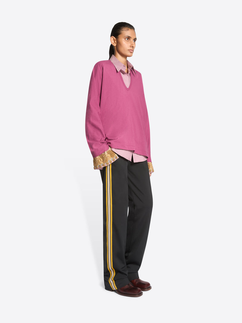 A model stands against a light gray background wearing a pink long-sleeve sweater over a light pink collared shirt, paired with dark gray track pants featuring yellow side stripes and dark brown shoes.