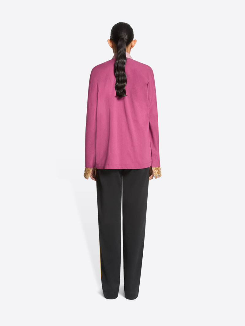 Back view of a model standing against a light gray background wearing a pink long-sleeve sweater over a light pink collared shirt, paired with dark gray track pants featuring yellow side stripes and dark brown shoes.