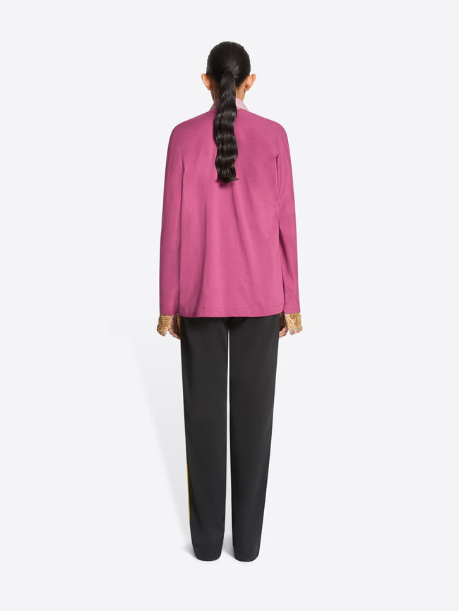 Back view of a model standing against a light gray background wearing a pink long-sleeve sweater over a light pink collared shirt, paired with dark gray track pants featuring yellow side stripes and dark brown shoes.